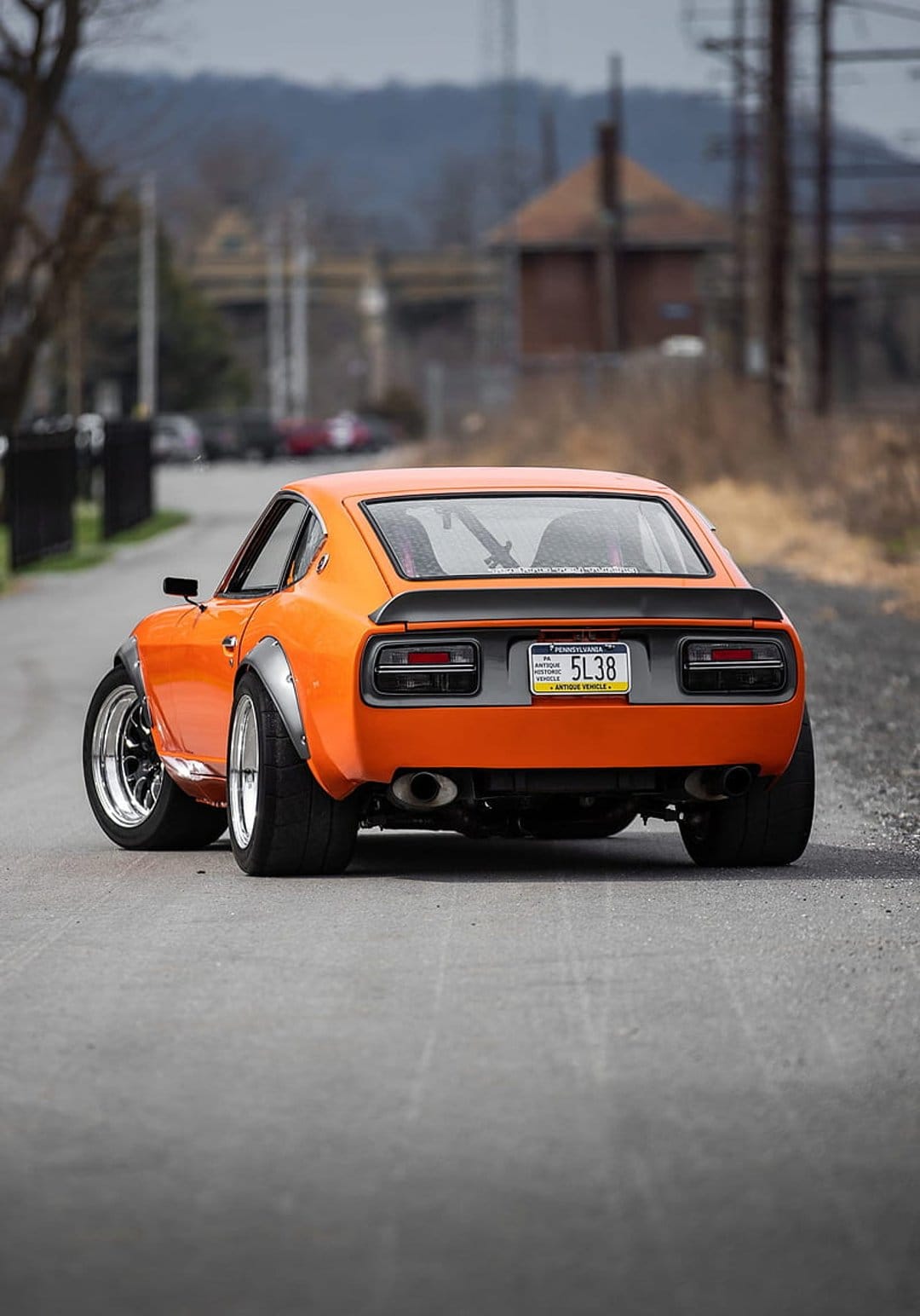 Orange Nissan 240Z with widebody fenders and a rear spoiler parked on a road, shown from a low rear perspective.