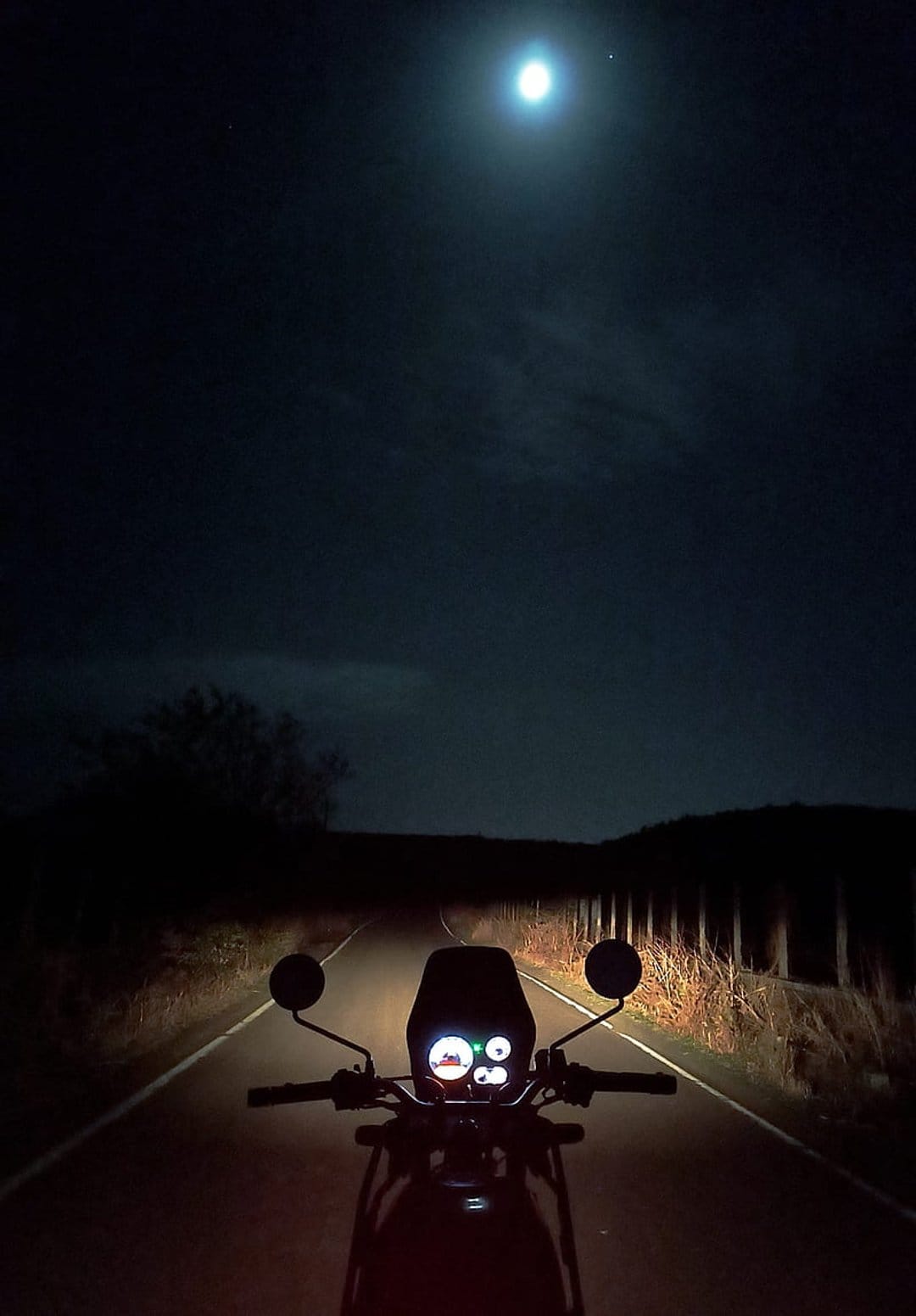 Motorcycle handlebars and glowing dashboard on a dark road under a large bright moon in a cloudy night sky.