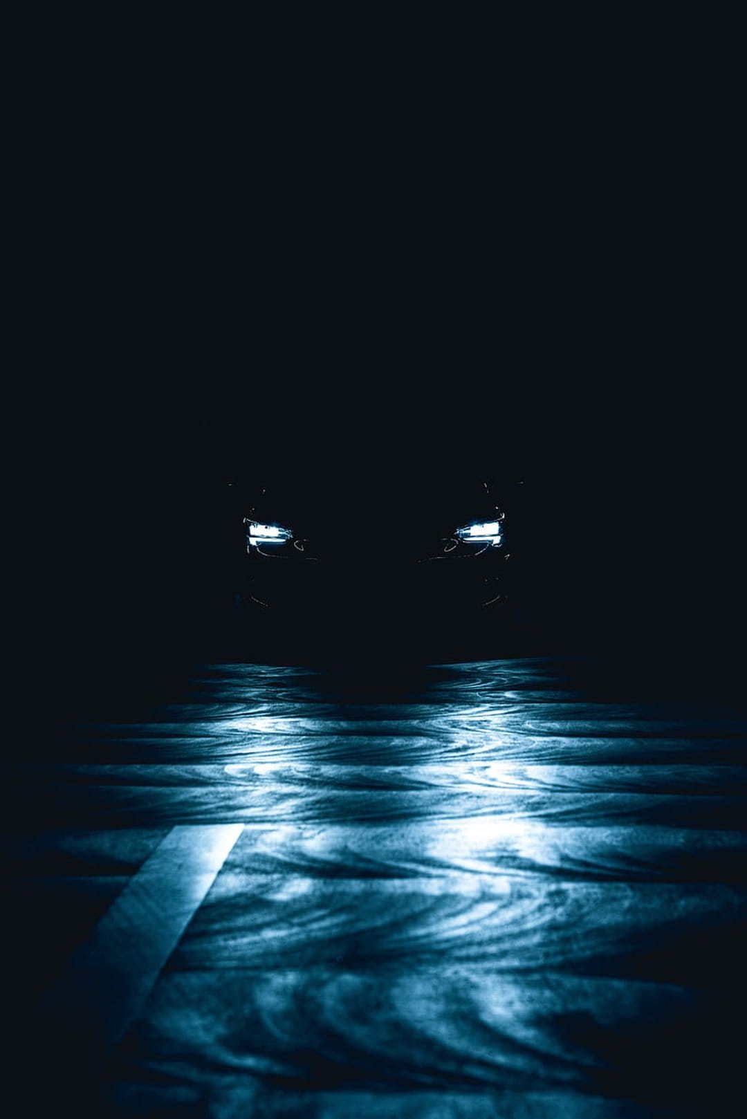 Blue car headlights reflect on a wet, textured road surface against a dark, nocturnal background.