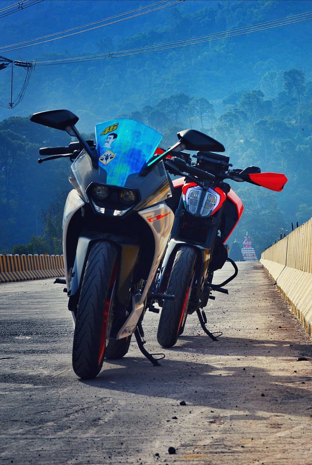 White and orange sportbike parked alongside a red motorcycle on a misty mountain road with a hazy blue background.
