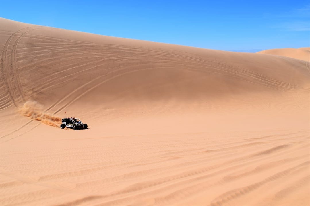 Dune buggy kicking up sand while racing over golden dunes under a bright blue sky with visible tire tracks.