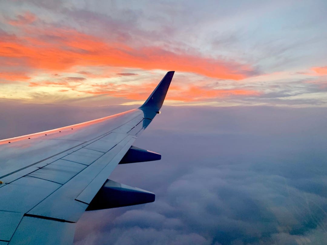Airplane wing extends over a thick layer of orange and pink clouds during a vibrant high-altitude sunset.