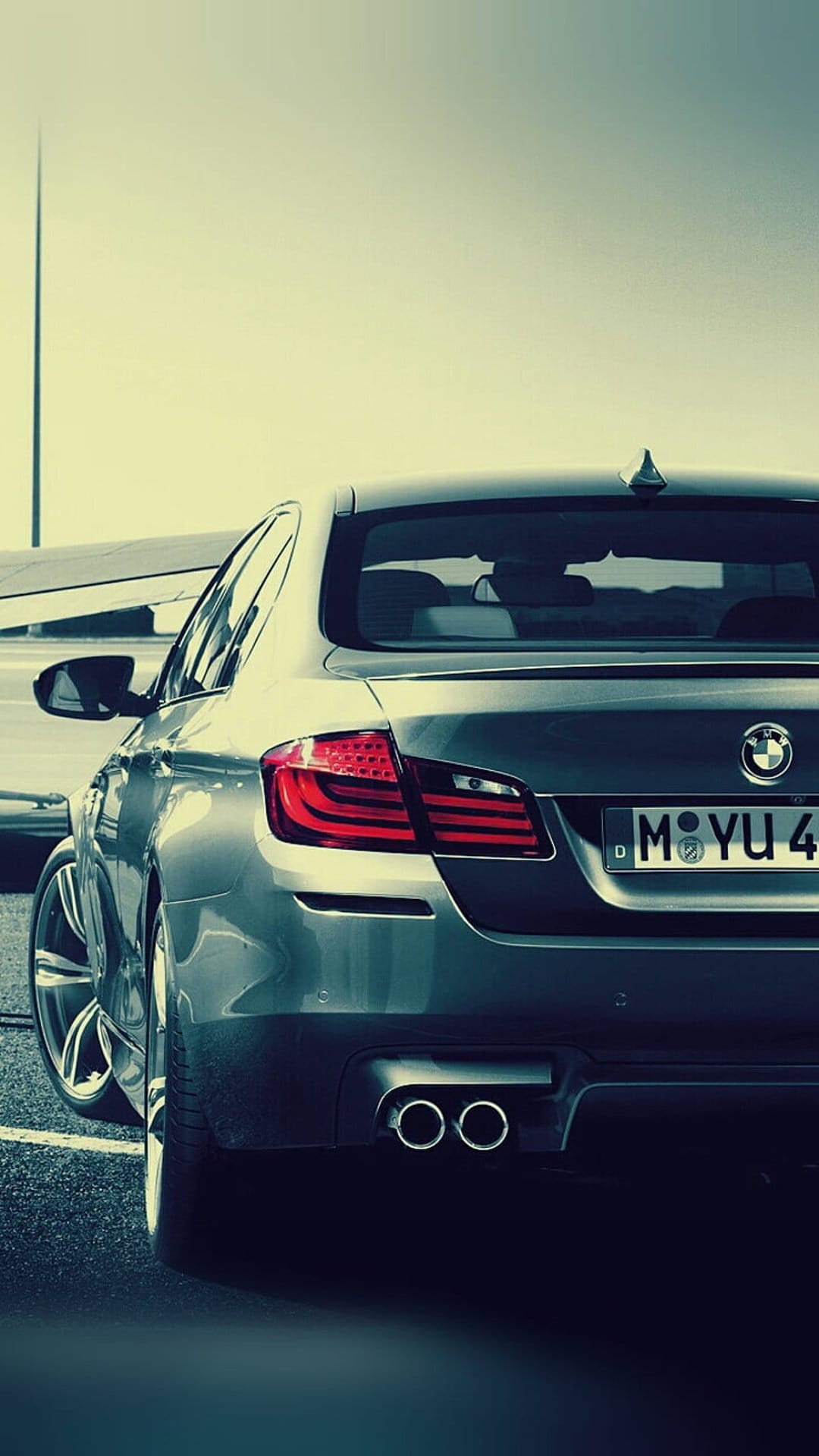 Silver BMW sedan rear view featuring red taillights and dual exhausts parked near a small airplane wing on asphalt.