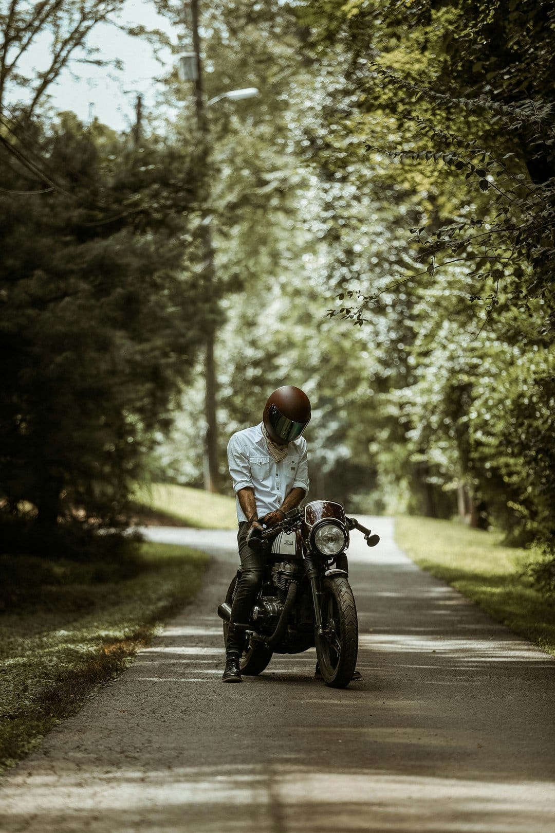 Biker in a white shirt sits on a retro motorcycle along a paved road lined with towering green deciduous trees.