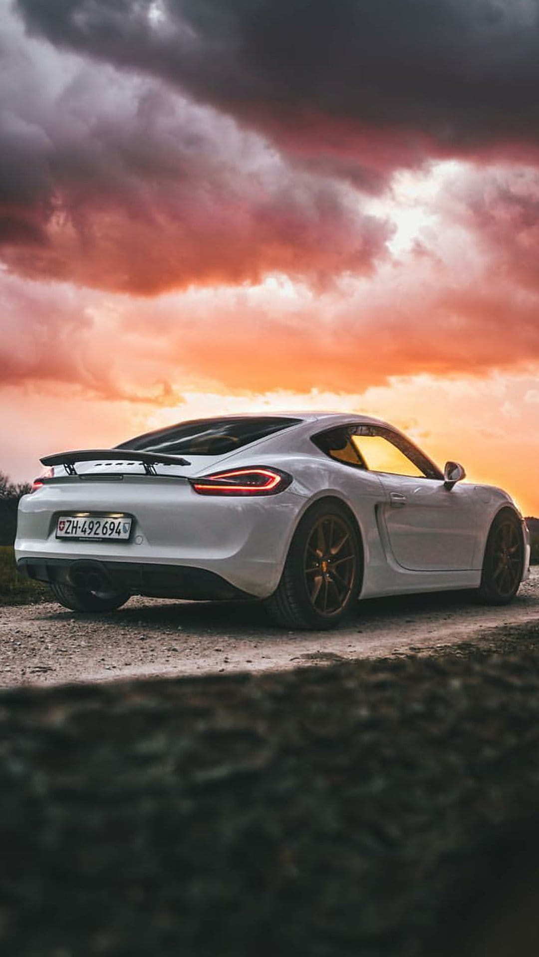 White Porsche Cayman with glowing taillights parked on a dirt road under dramatic orange and dark clouds.