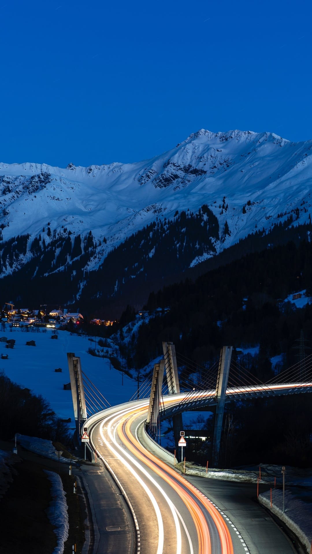 Luminous light trails stretch across a modern bridge toward snow-covered mountains under a dark twilight sky.