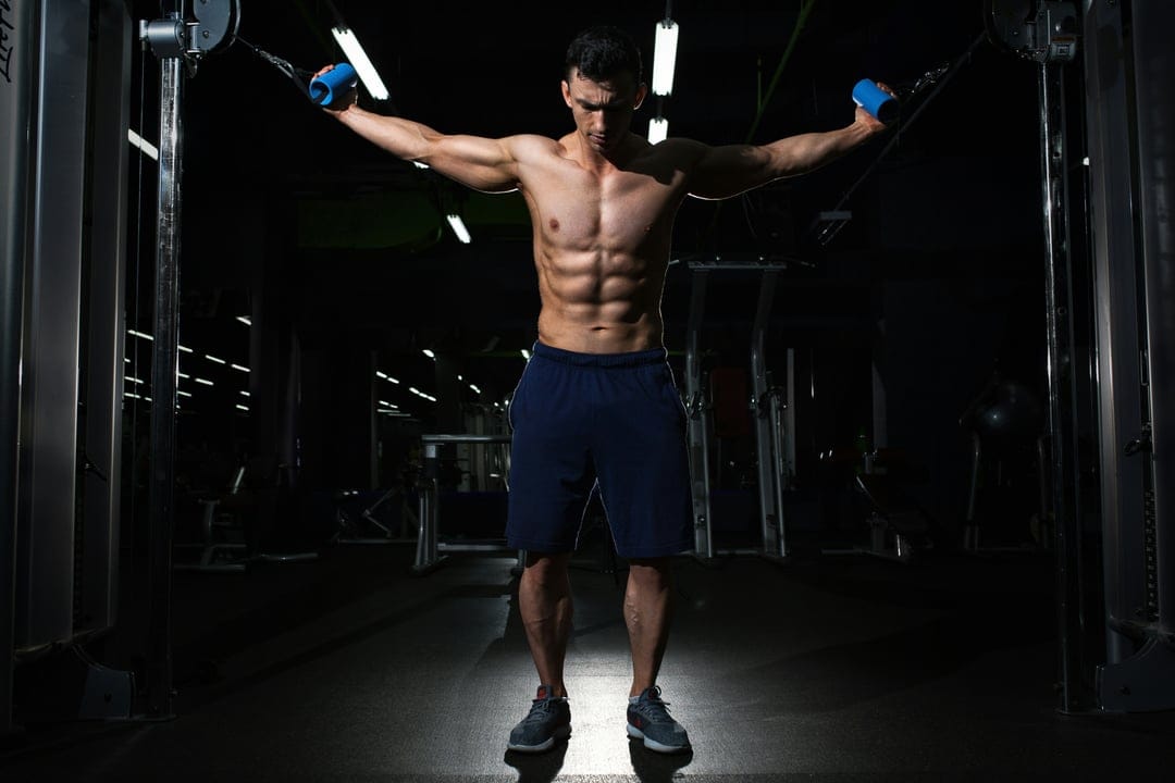Muscular man performing cable fly exercises with handles in a dark modern gym, showing defined abdominal muscles.