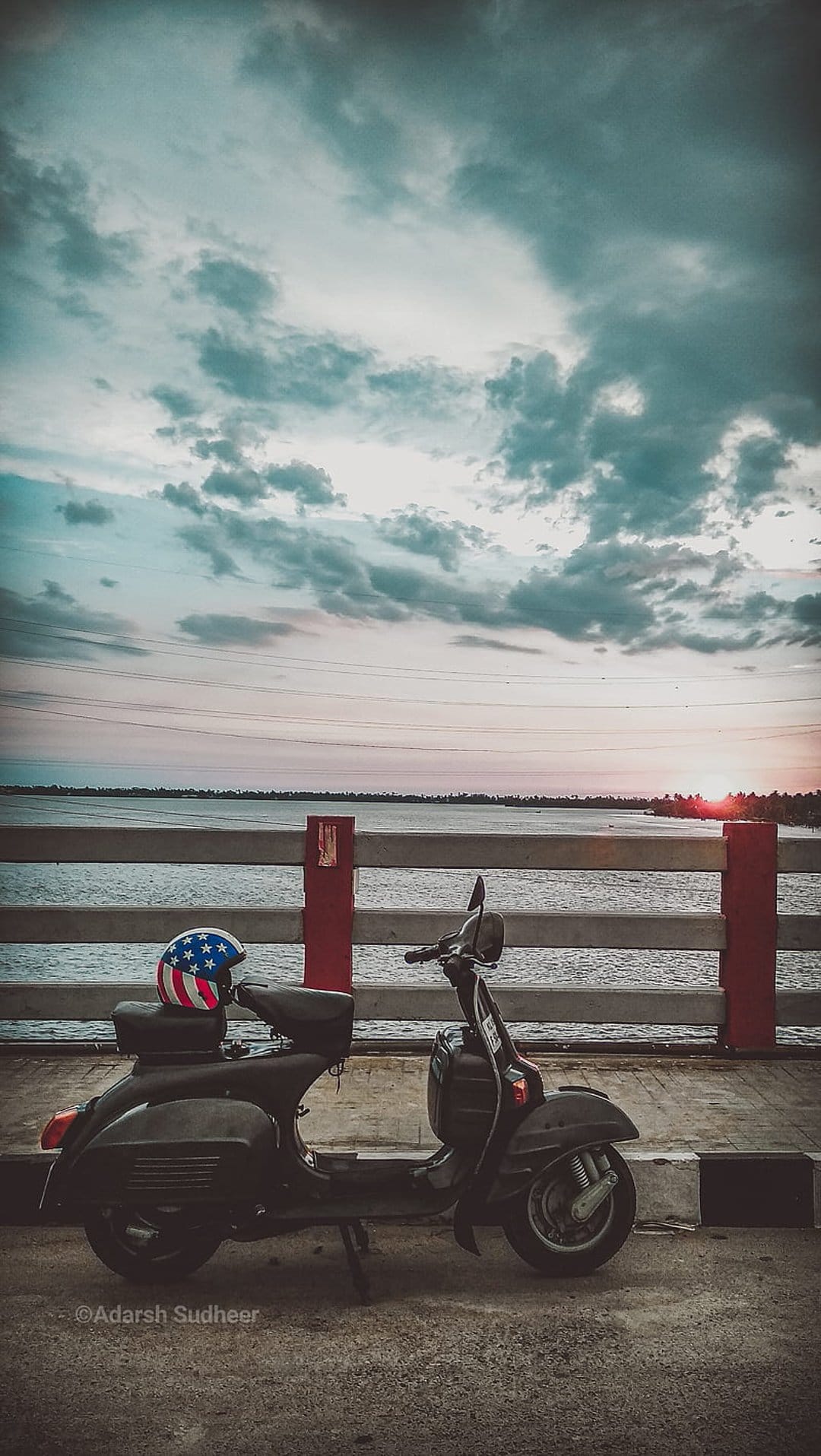 Vintage black scooter and American flag helmet on a bridge during a golden sunset with dramatic clouds over water.