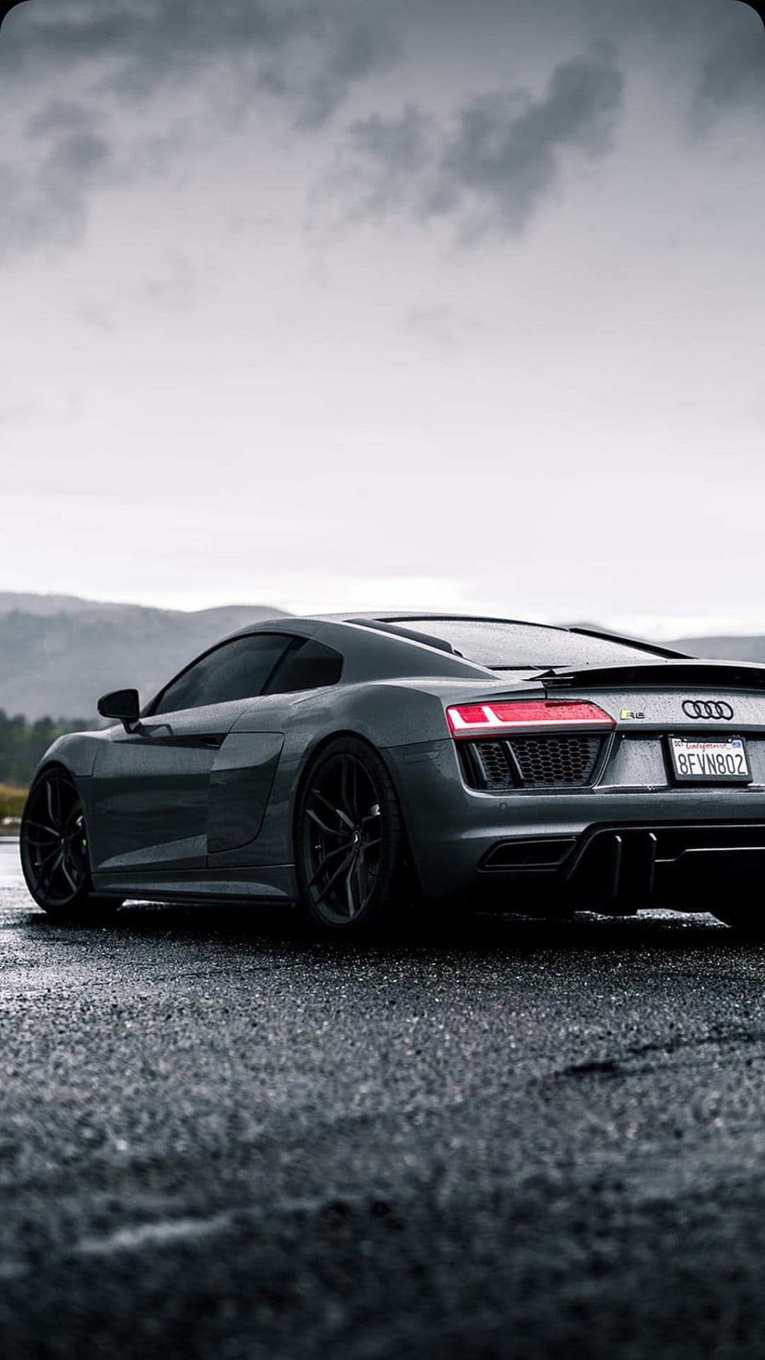 Audi R8 rear view with glowing red taillights reflecting on a wet asphalt road against misty mountain peaks.