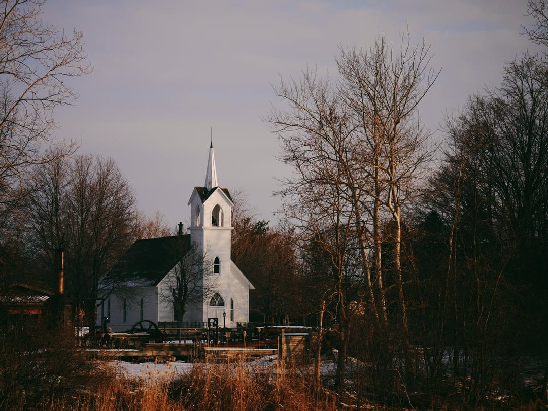White church steeple rises behind dark, leafless tree branches on a serene and snow-dusted winter landscape.