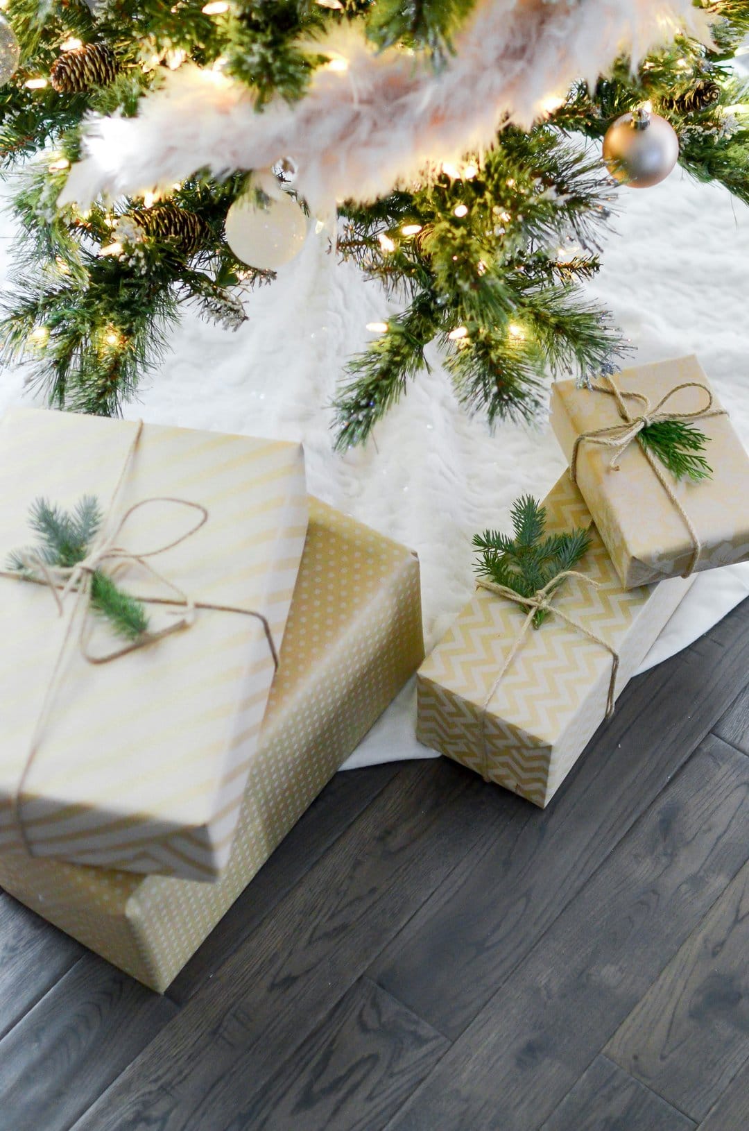 Christmas gifts in kraft and gold paper rest on white faux fur beneath decorated evergreen pine branches.