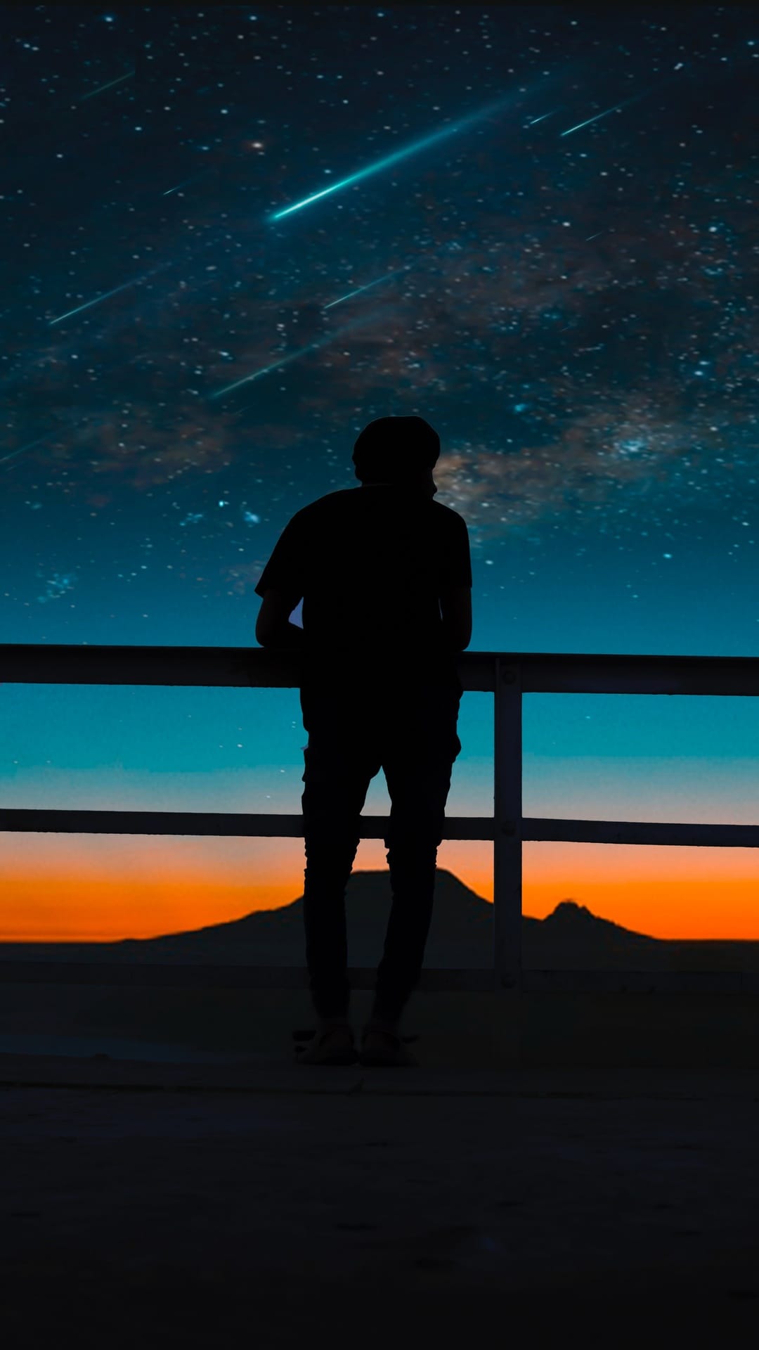 Person standing on a balcony watching a purple galaxy sky with shooting stars over distant mountain peaks.