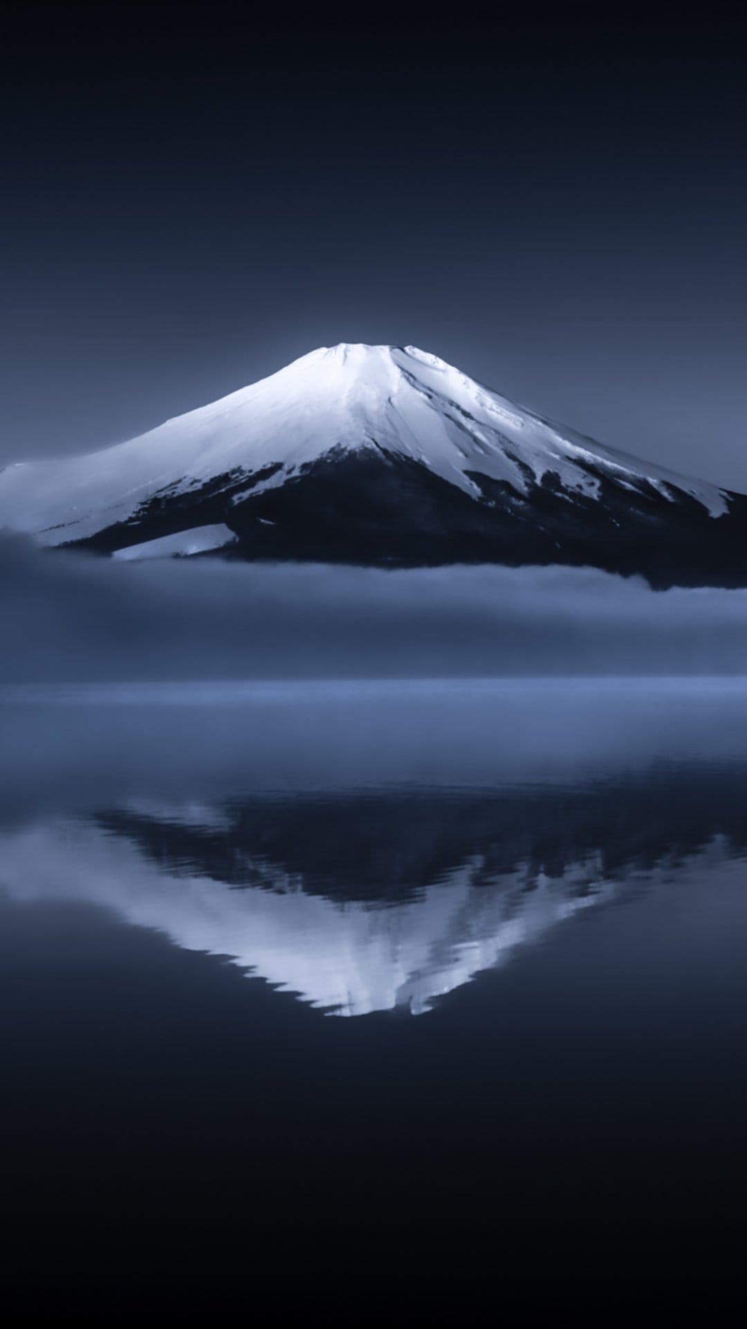 Mount Fuji reflected in dark water beneath a foggy sky with a snow-covered summit and deep monochromatic tones.