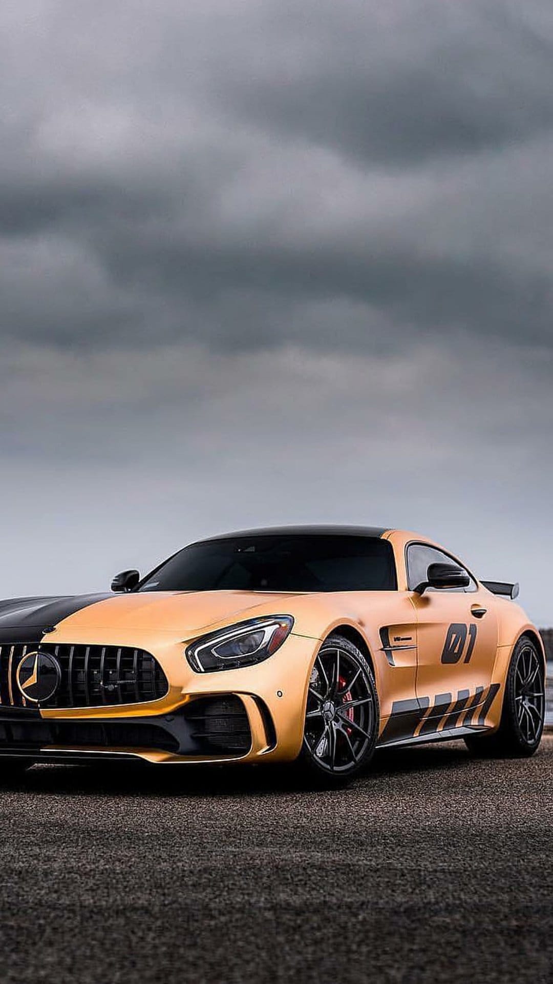 Golden Mercedes-AMG GT R with a black racing stripe and 01 side decal parked on dark asphalt under cloudy skies.
