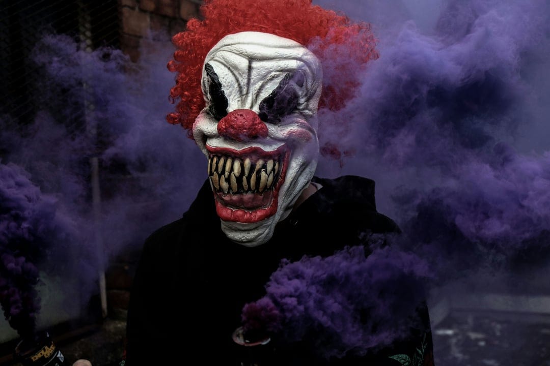 Masked clown with sharp teeth holding two purple smoke bombs that emit thick plumes of violet vapor against a dark backdrop.