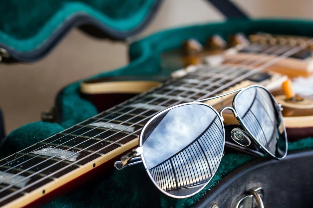 Aviator sunglasses reflecting a cloudy sky rest on the strings of a guitar nestled in a green velvet case.