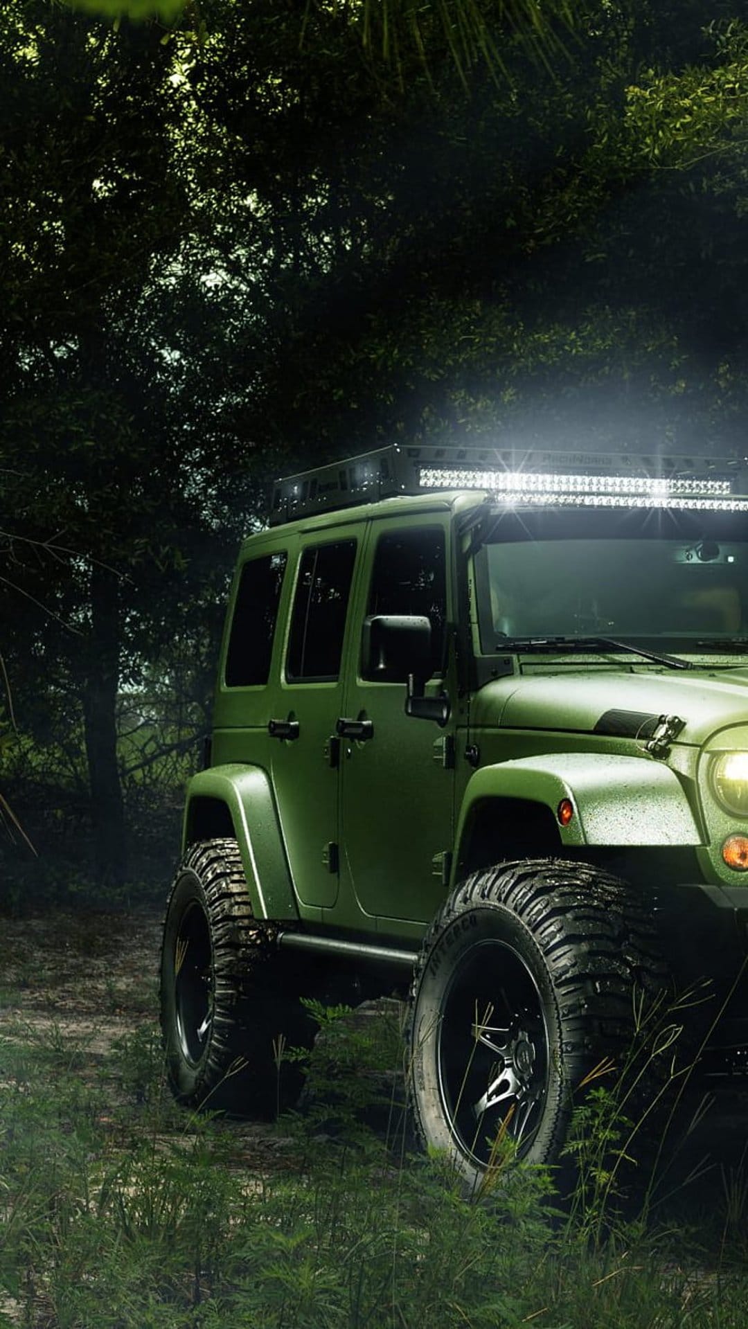 Olive green Jeep Wrangler with a lit LED bar parked on a woodland trail, highlighting aggressive tires and trees.