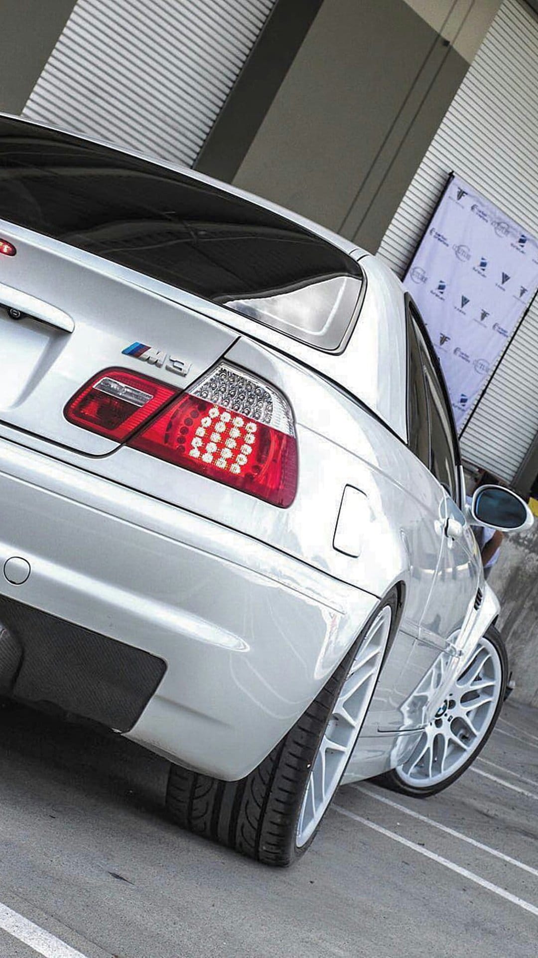Silver BMW M3 rear with glowing red LED tail lights and white multi-spoke wheels on a textured background.