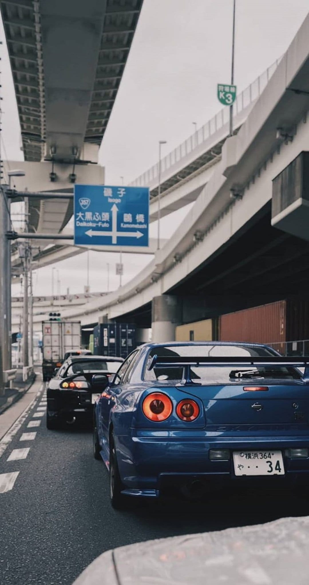Blue Nissan Skyline and black Toyota Supra drive on a Tokyo highway with shipping containers in the background.