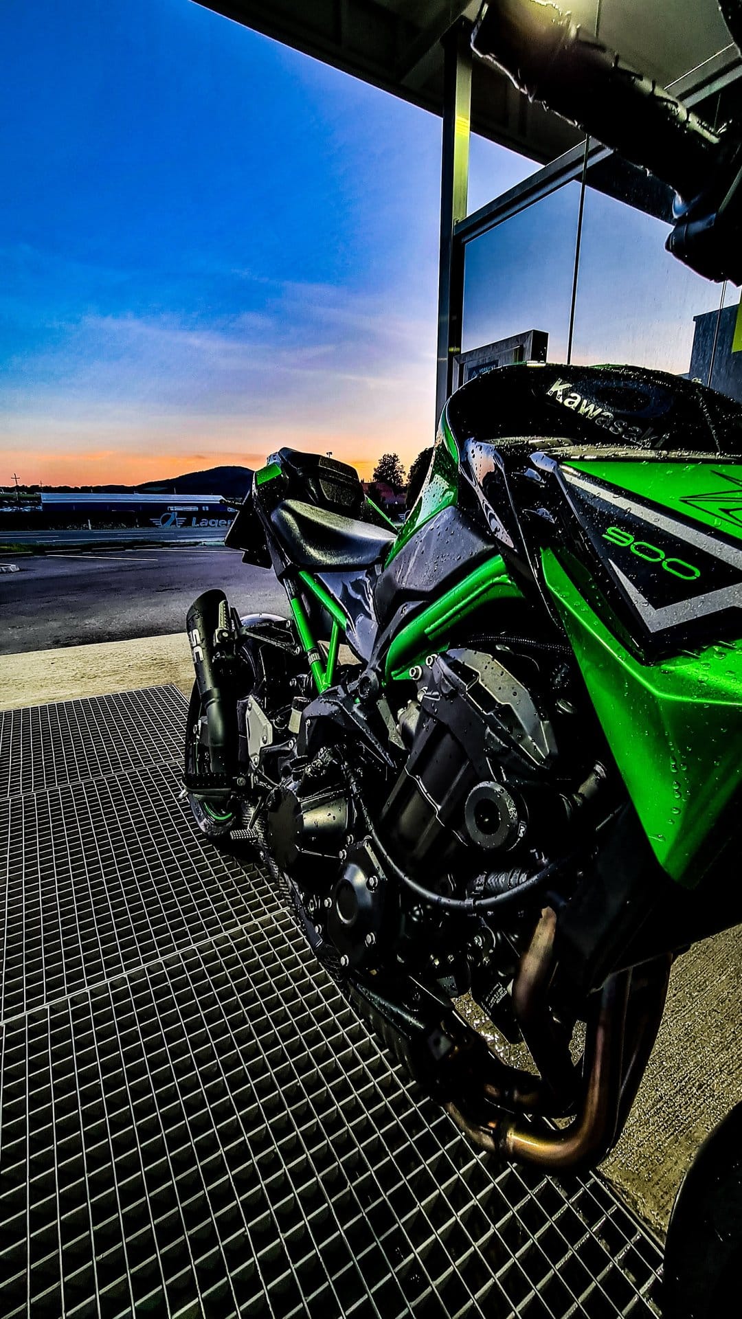 Green Kawasaki Z900 motorcycle on a metal grate floor against a vibrant orange and purple sunset sky.