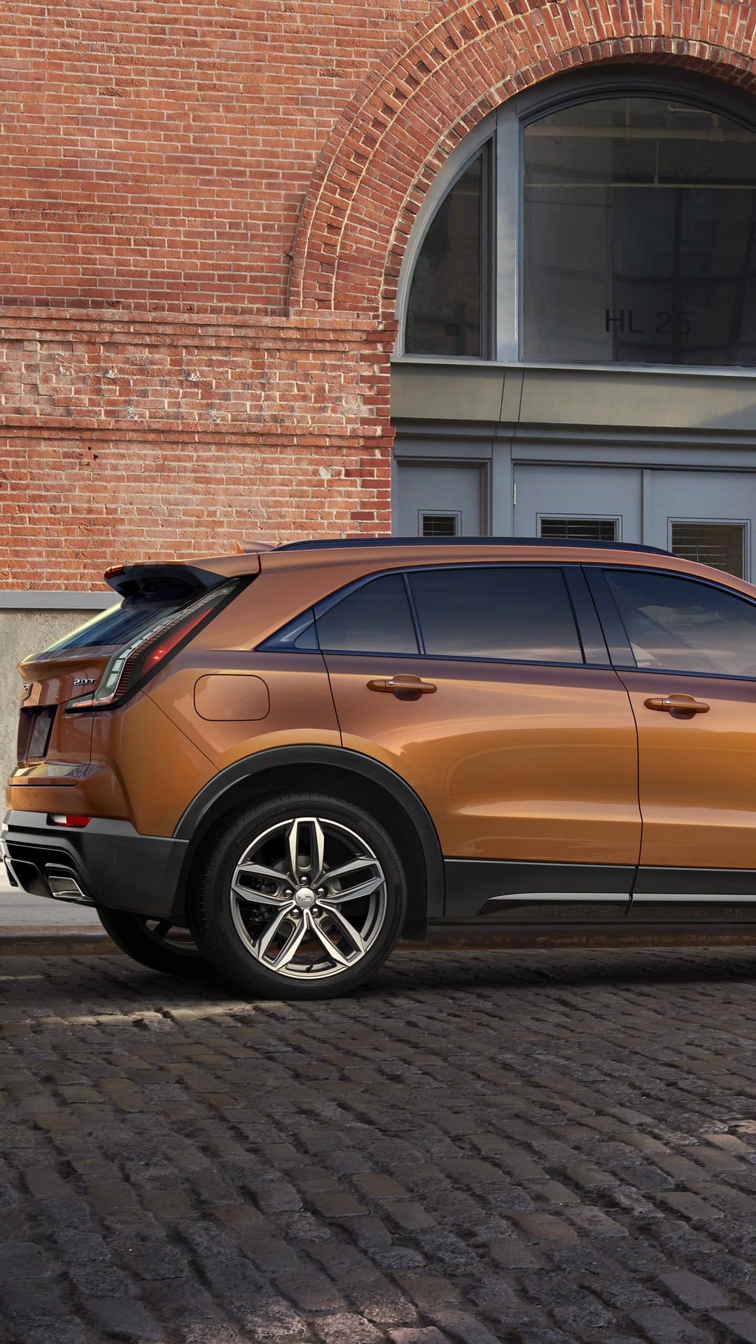 Copper Cadillac XT4 parked on a cobblestone street against a red brick building with a large arched window.