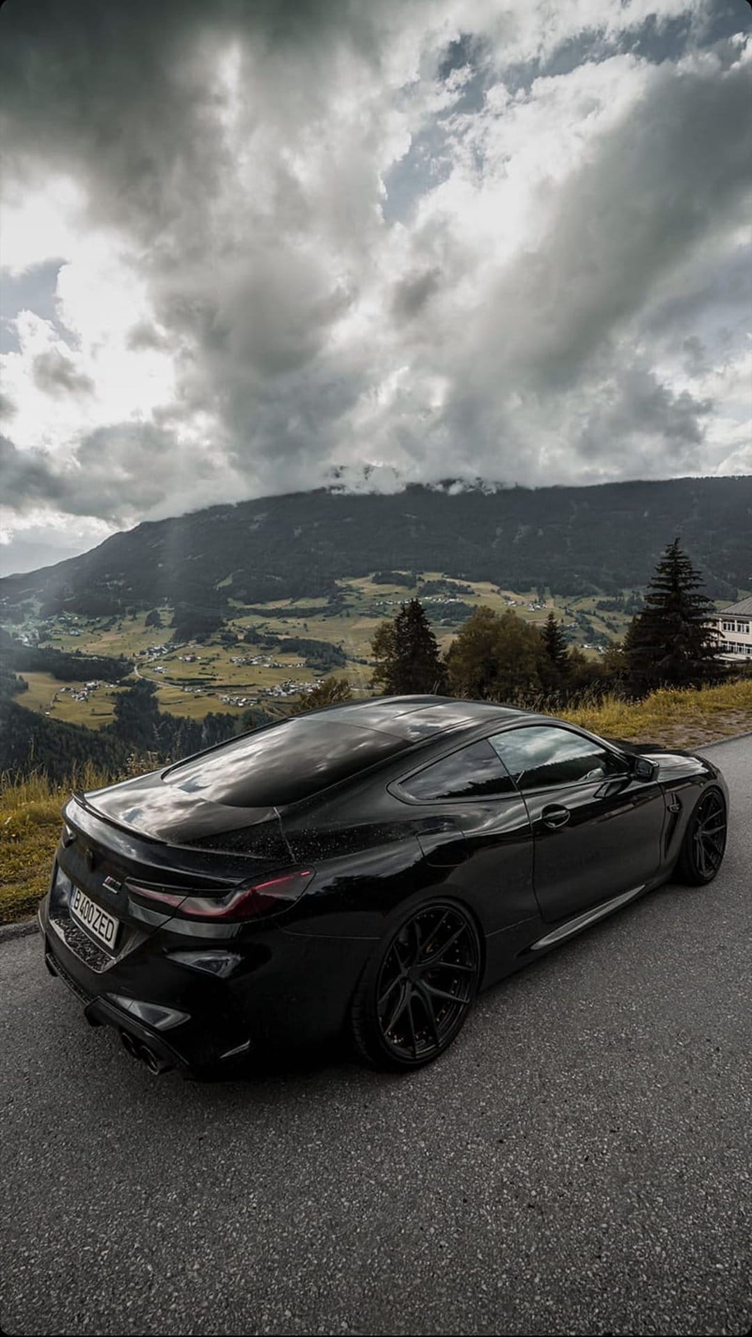 Black BMW coupe parked on a paved mountain road under a dark, cloudy sky with jagged alpine peaks in the distance.