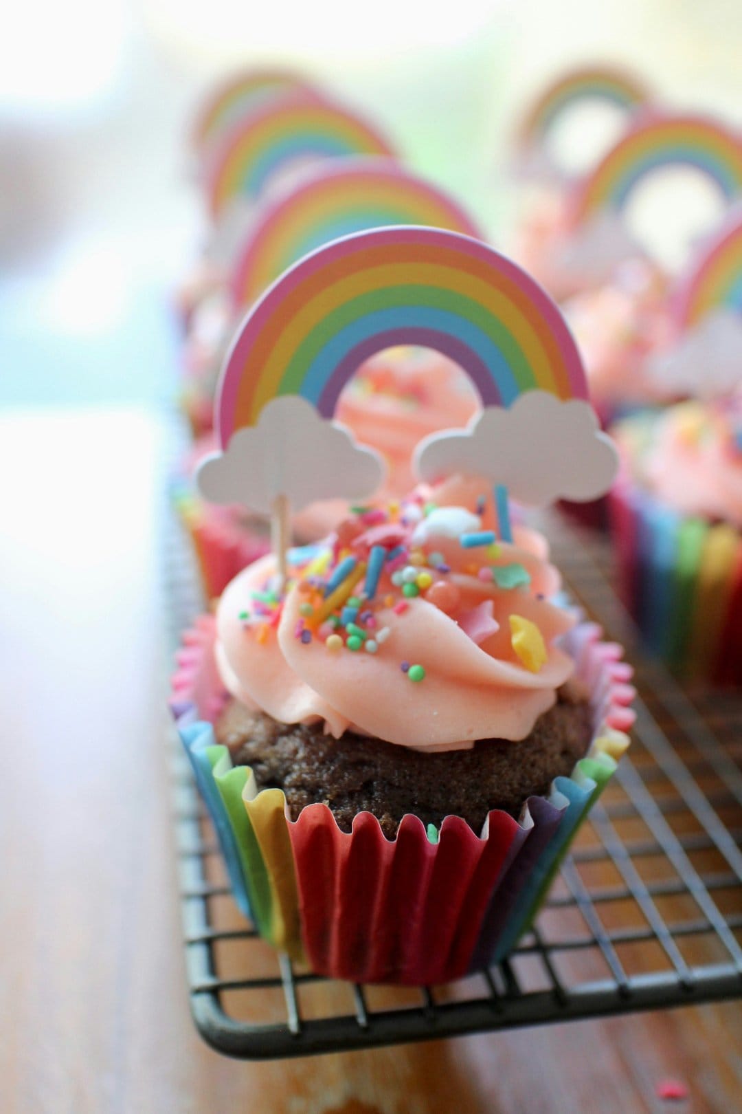 Pink buttercream cupcakes with rainbow toppers and colorful sprinkles are lined up on a metal cooling rack.
