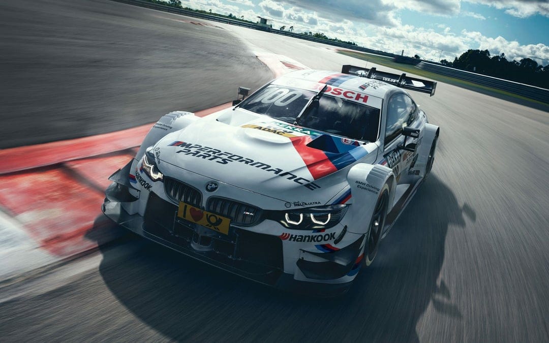White BMW M4 DTM race car with red and blue stripes cornering fast on a racetrack with motion blur.