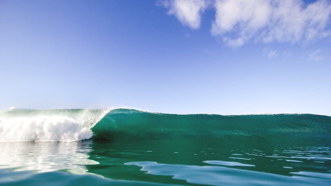 Turquoise barrel wave curling over clear water with white sea foam under a blue sky with scattered clouds.