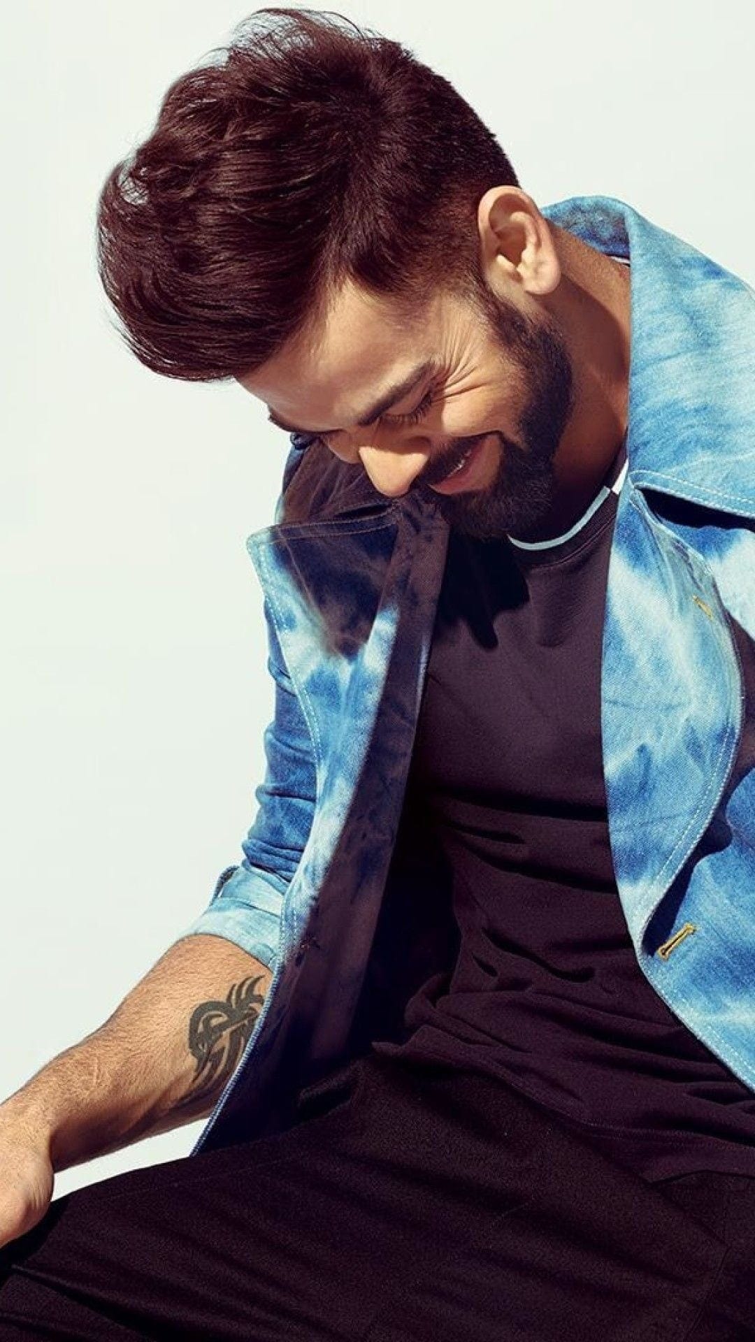 Virat Kohli laughs with head bowed wearing a tie-dye denim jacket and showing a detailed forearm tattoo.