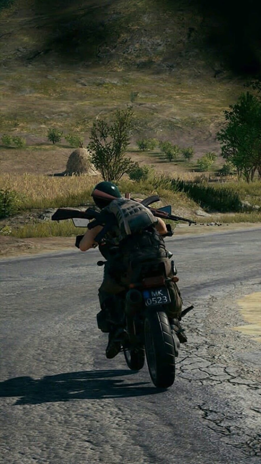 Soldier in tactical gear holding a rifle while riding a motorcycle on a cracked asphalt road in the countryside.