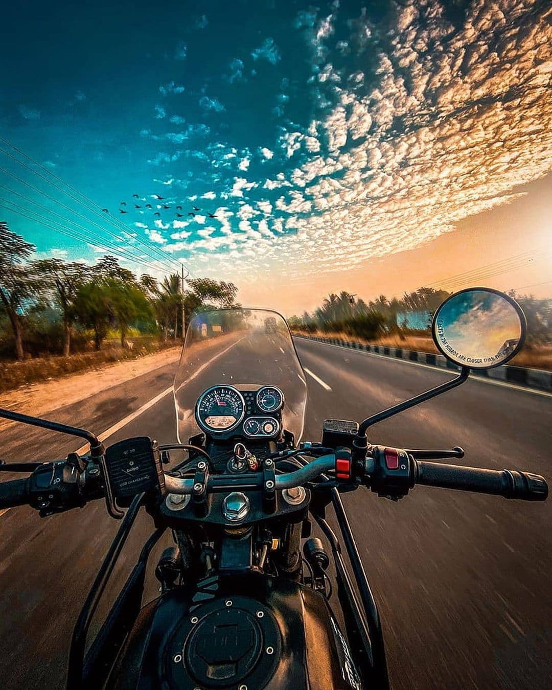 Motorcycle handlebars and illuminated gauges face a winding road under a vibrant orange and blue sunset sky.