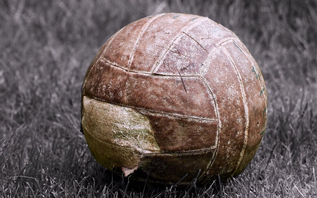 Weathered vintage leather soccer ball with worn stitching resting on textured greyish-green grass in a park.