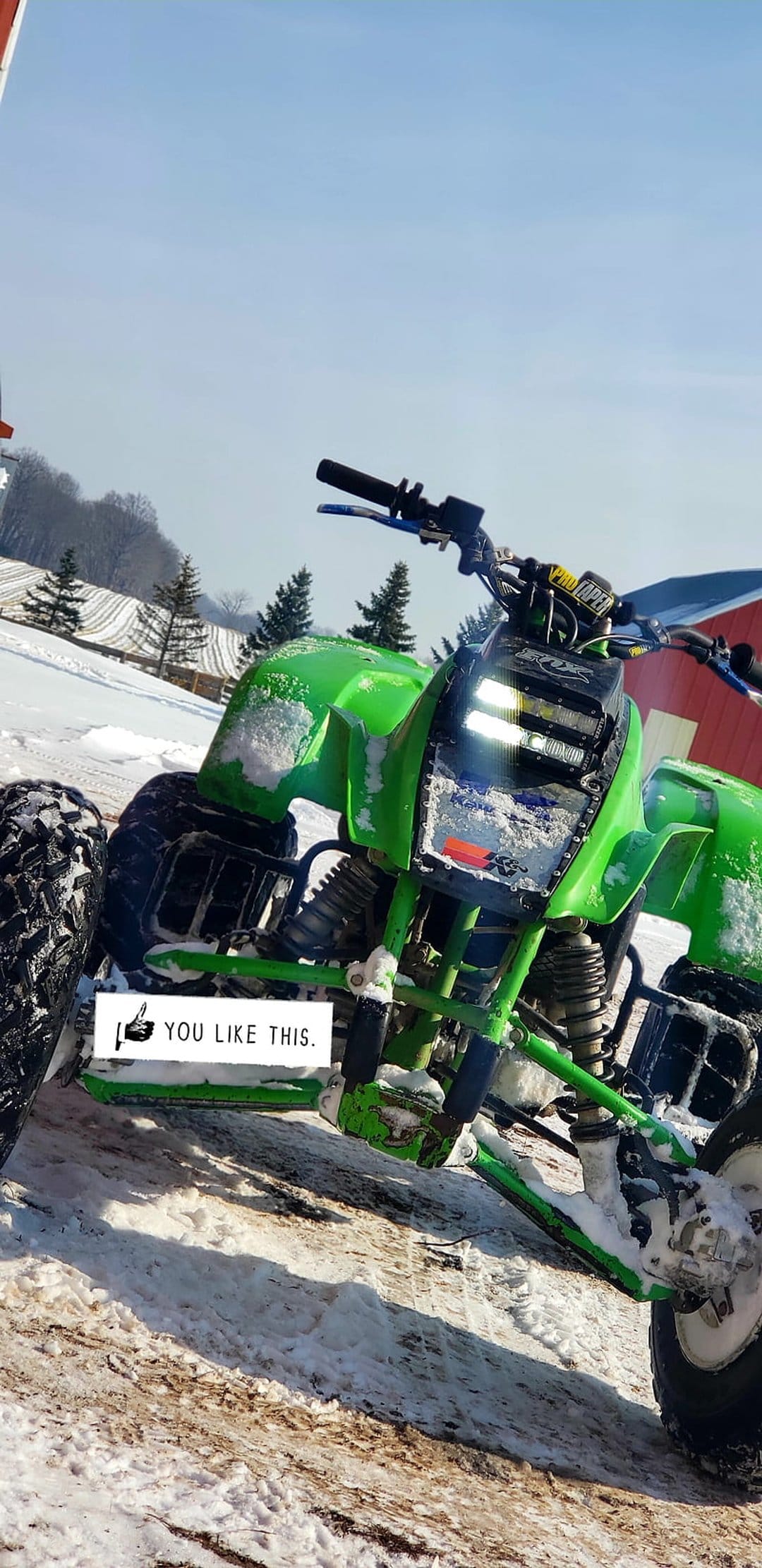 Green ATV with glowing headlights stands in a snowy field near a red barn and trees in a rural winter scene.