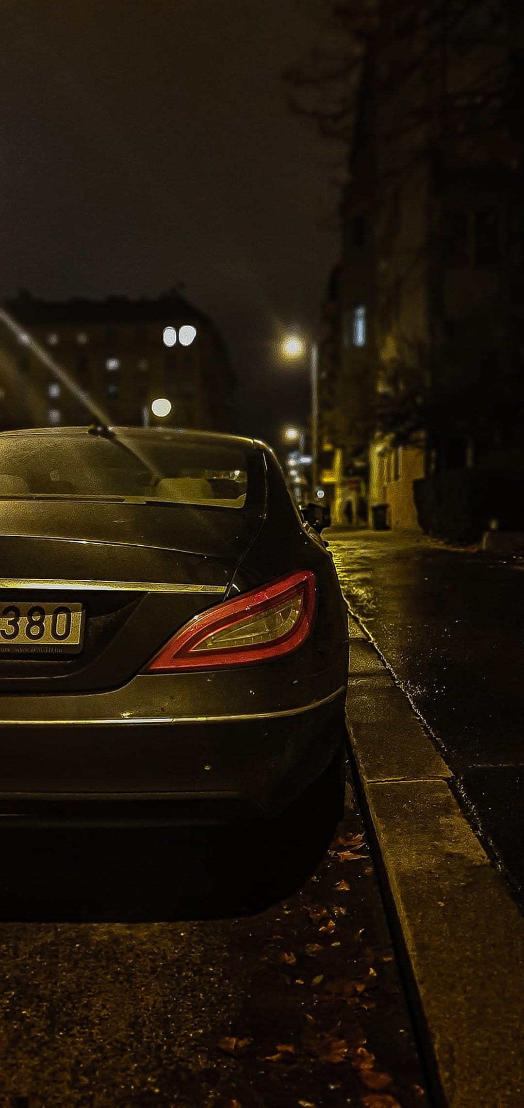 Car taillight glows red over a wet asphalt street with reflections of city lights in a vertical night scene.