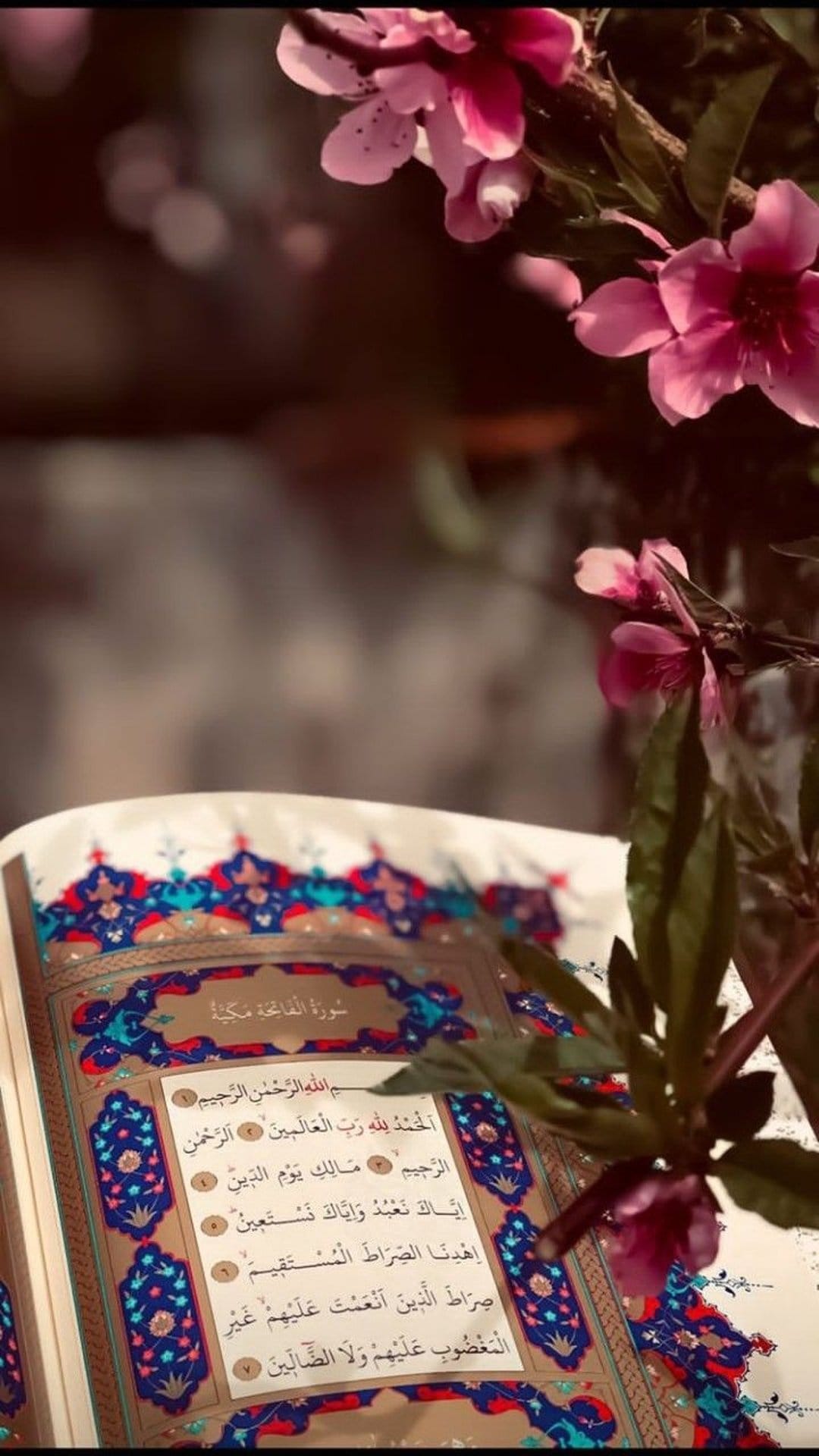 Open Quran with intricate Arabic calligraphy rests beside soft pink flowers in a bright, tranquil setting.