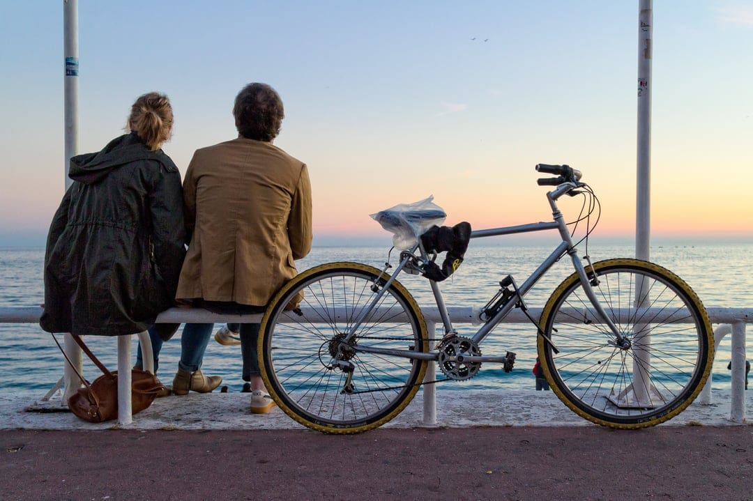 Two people sit on a bench with a bicycle nearby, overlooking a vibrant orange and pink sunset above calm sea water.