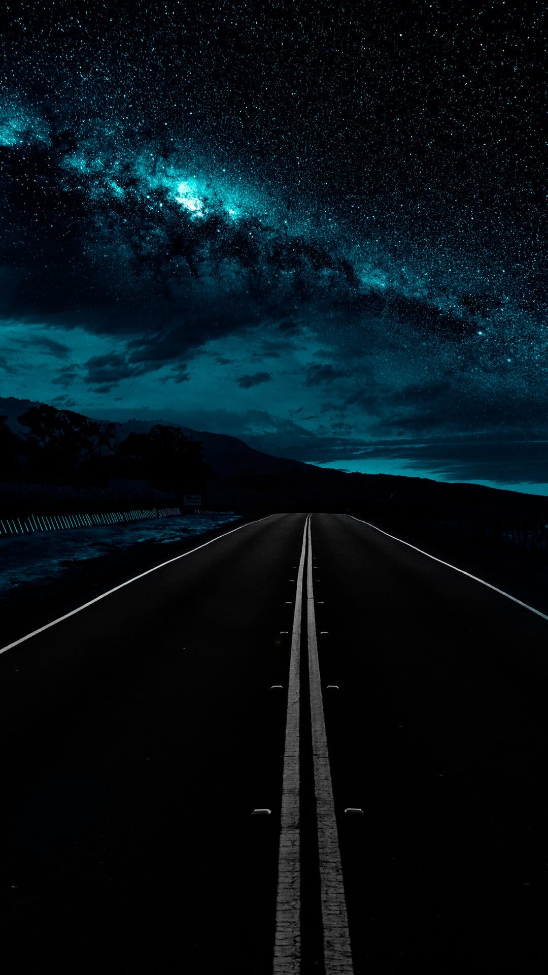 Asphalt road stretching into a luminous blue galaxy with silhouettes of mountains under a starry night.