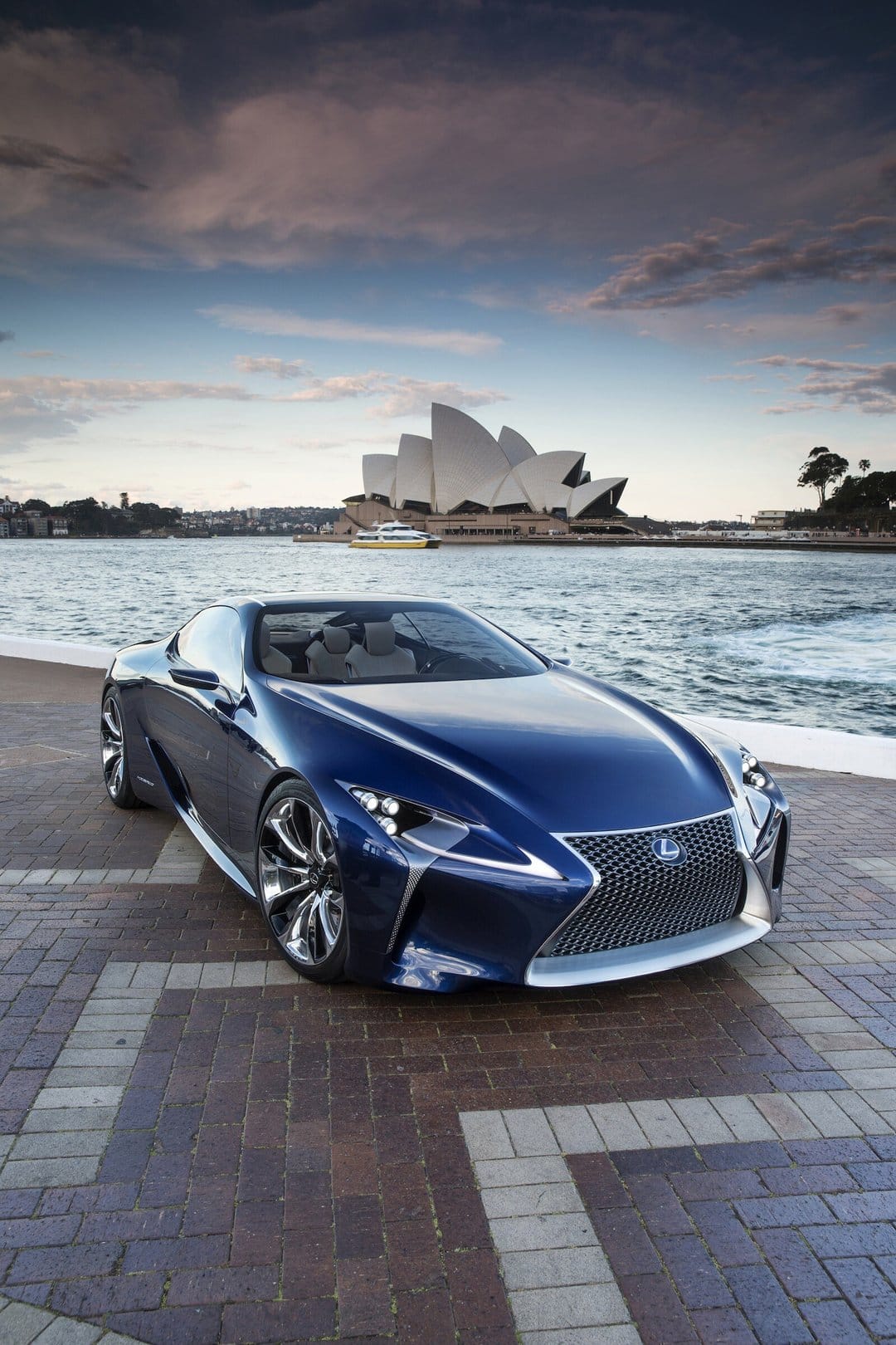 Blue luxury concept car parked on a brick pier with the Sydney Opera House and harbor in the background at dusk.