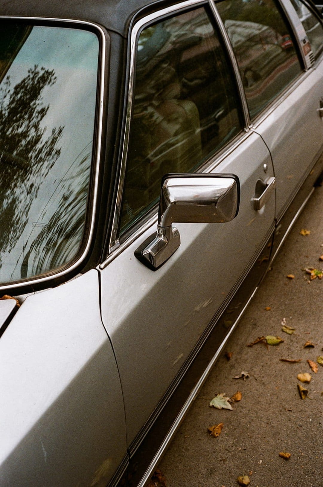 Vintage silver car door and chrome side mirror reflecting buildings above scattered autumn leaves on the ground.