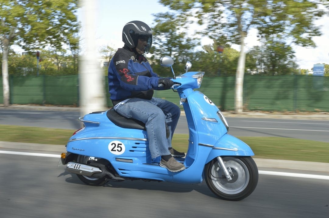 Man riding a blue Peugeot Django scooter with number 25 on a road with blurred background trees.