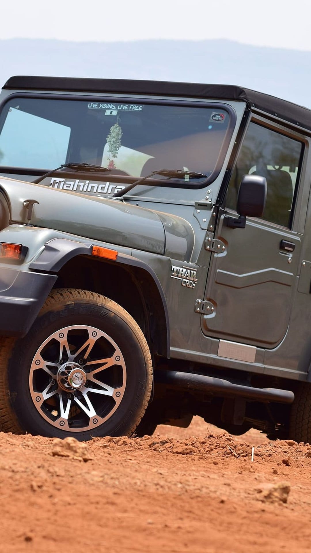 Grey Mahindra Thar SUV parked on reddish-brown dirt showing the front fender, tire, and windshield text.