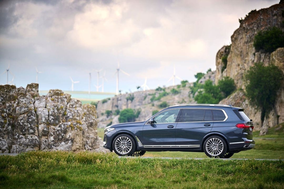 Dark blue BMW X7 SUV parked on a roadside with rocky cliffs and wind turbines under a cloudy sky.