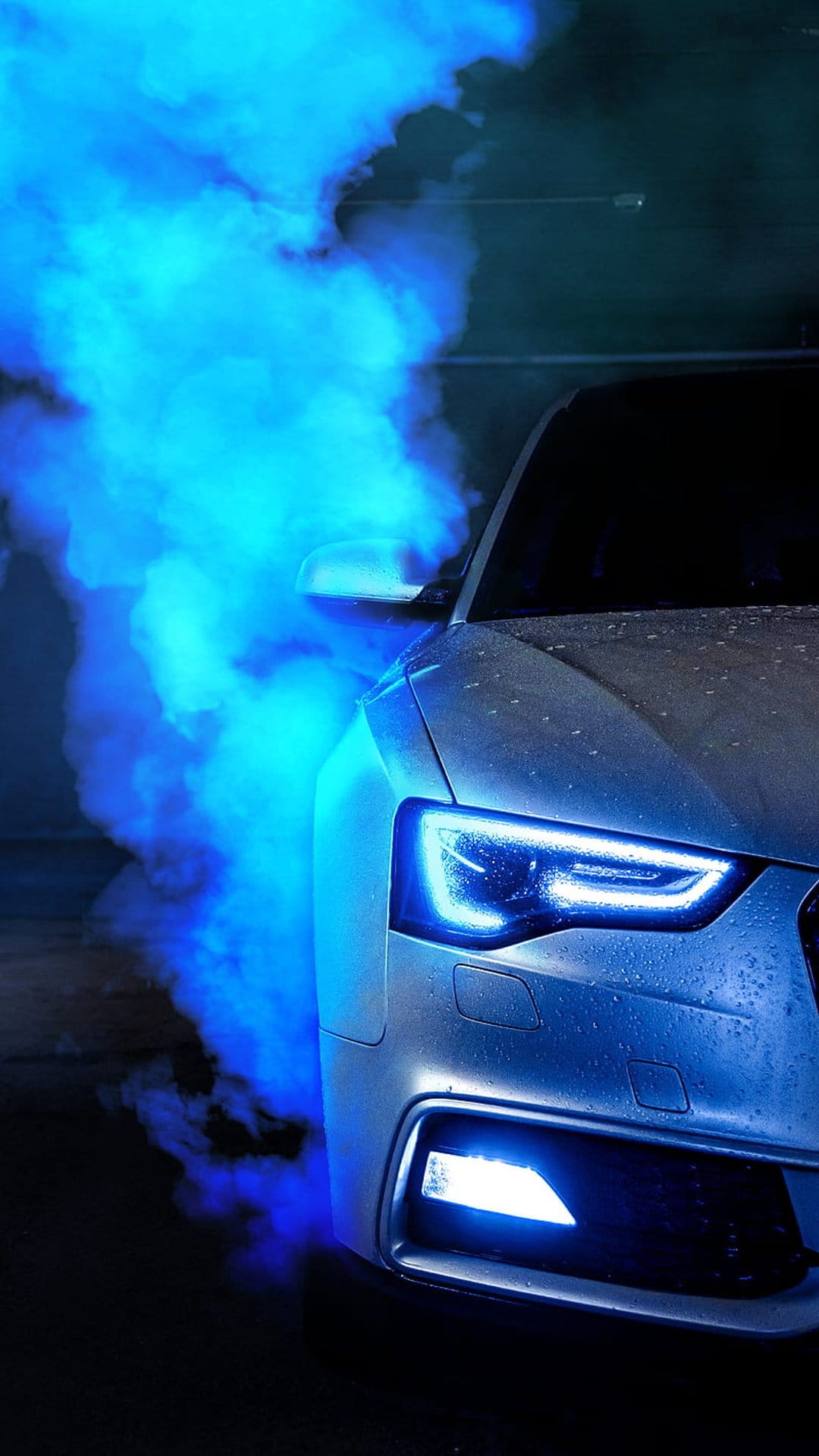 Silver Audi car front end with glowing LED headlights partially obscured by thick clouds of vibrant blue smoke.