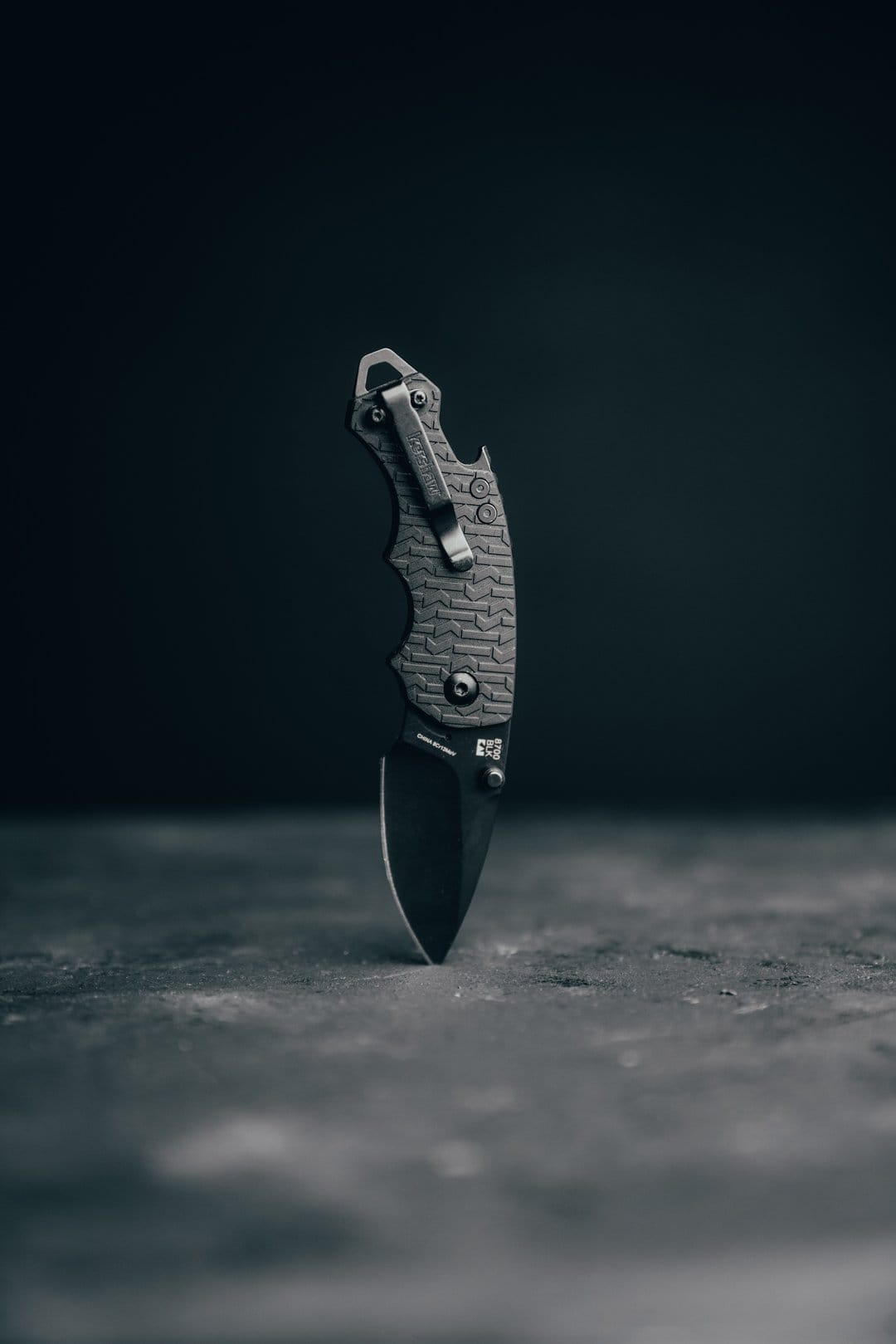 Black folding knife with a textured handle and pocket clip stands upright on a dark, gritty studio surface.