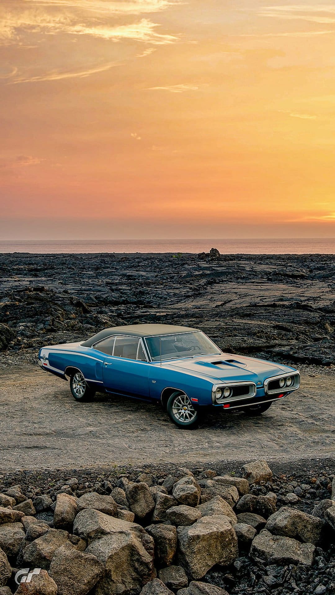 Blue Dodge Coronet muscle car parked on a rocky coast under a vibrant orange and pink sunset sky.