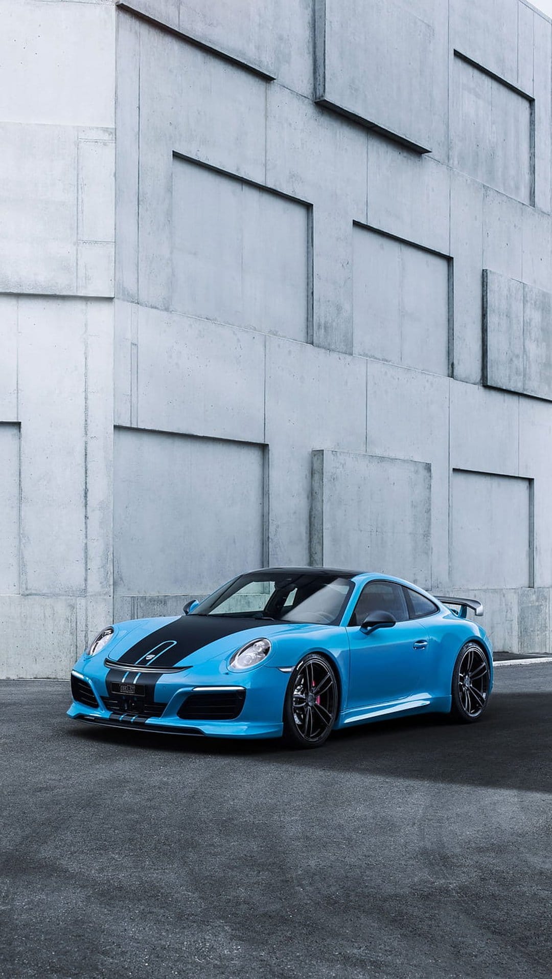 Blue Porsche 911 sports car with black racing stripes parked on a concrete floor before a gray brutalist wall.