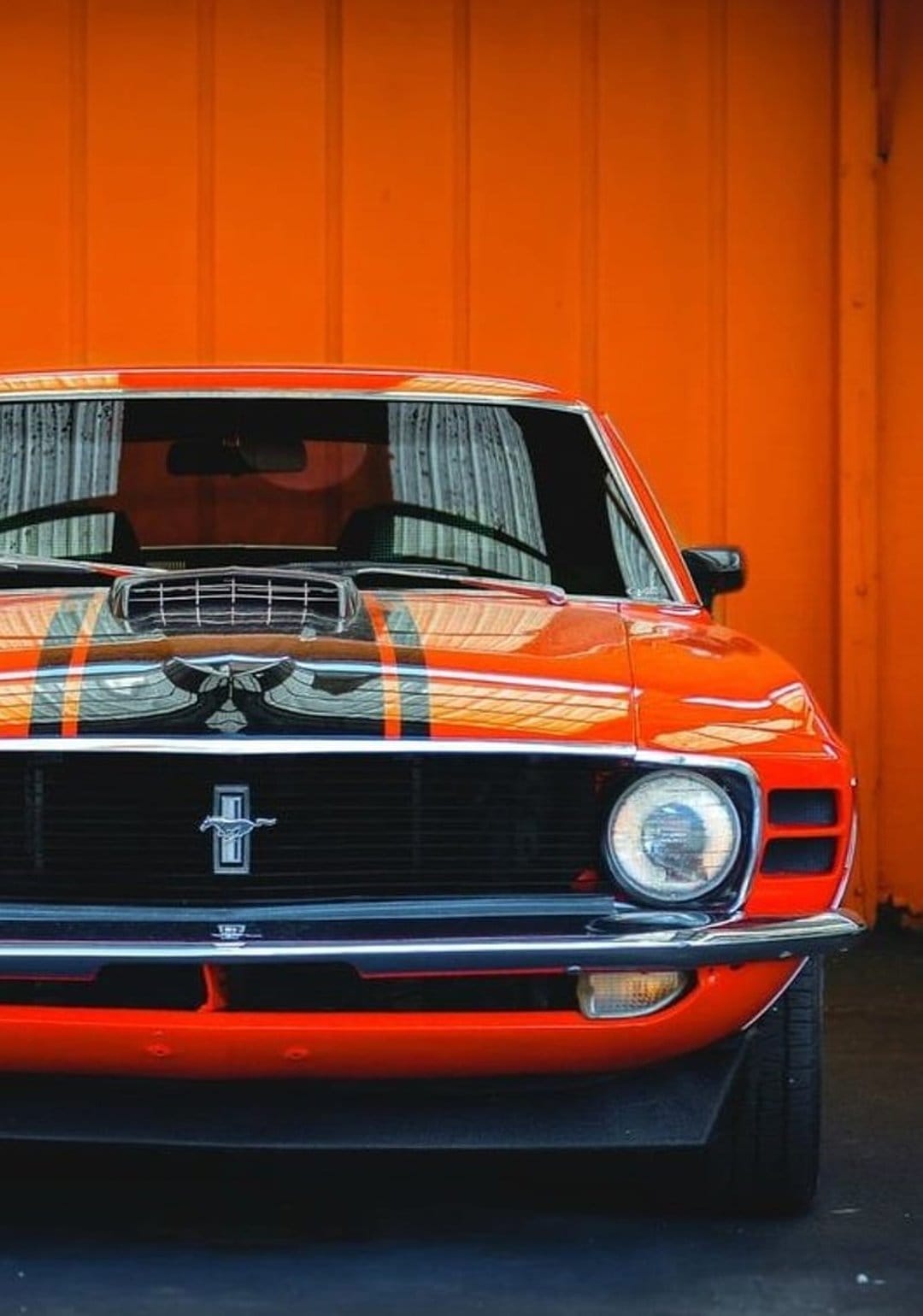 Classic orange Ford Mustang with black racing stripes parked against a matching textured orange vertical wall.