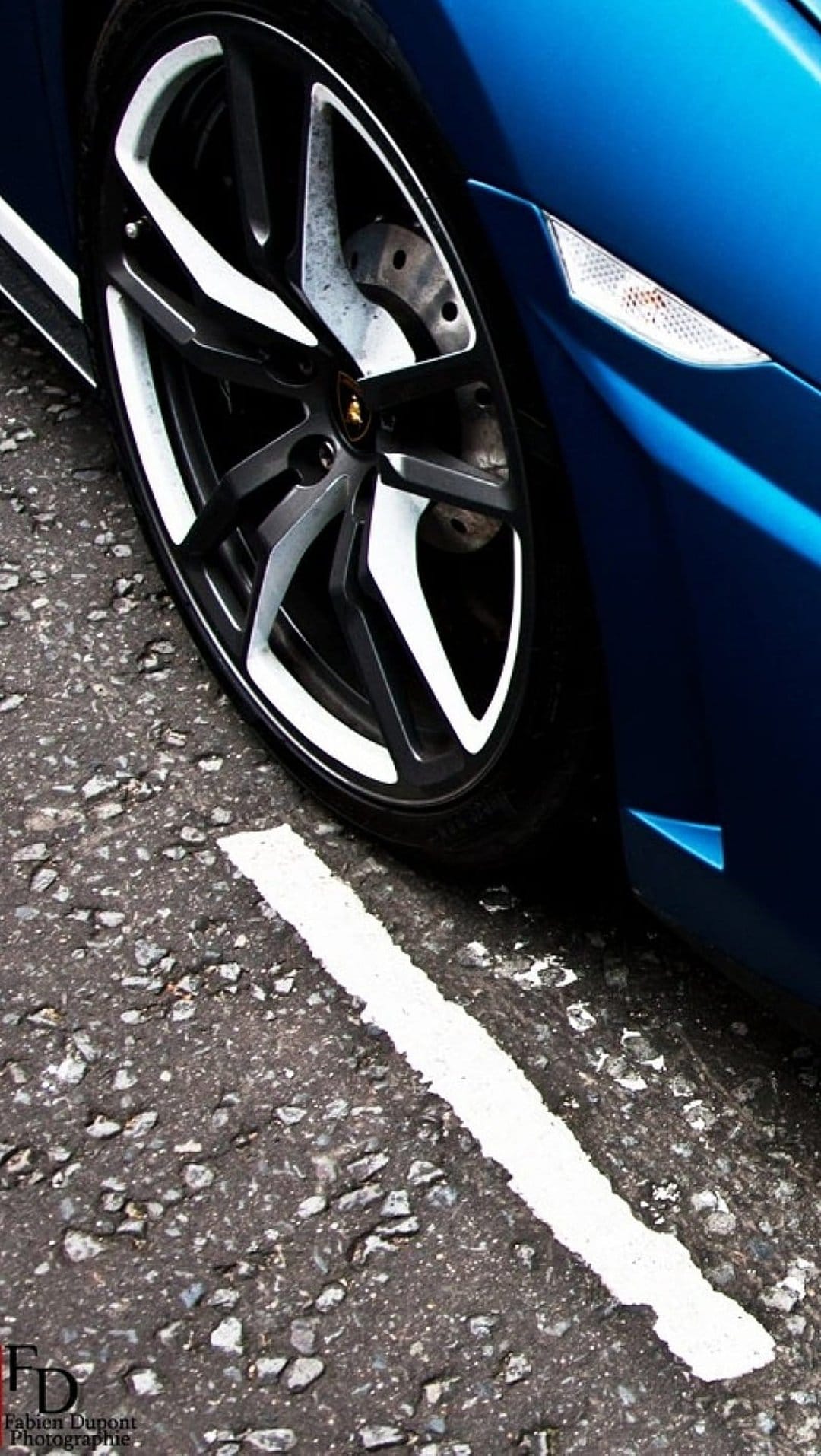 Blue Lamborghini alloy wheel with black and white spokes sits on dark asphalt next to a crisp white road stripe.