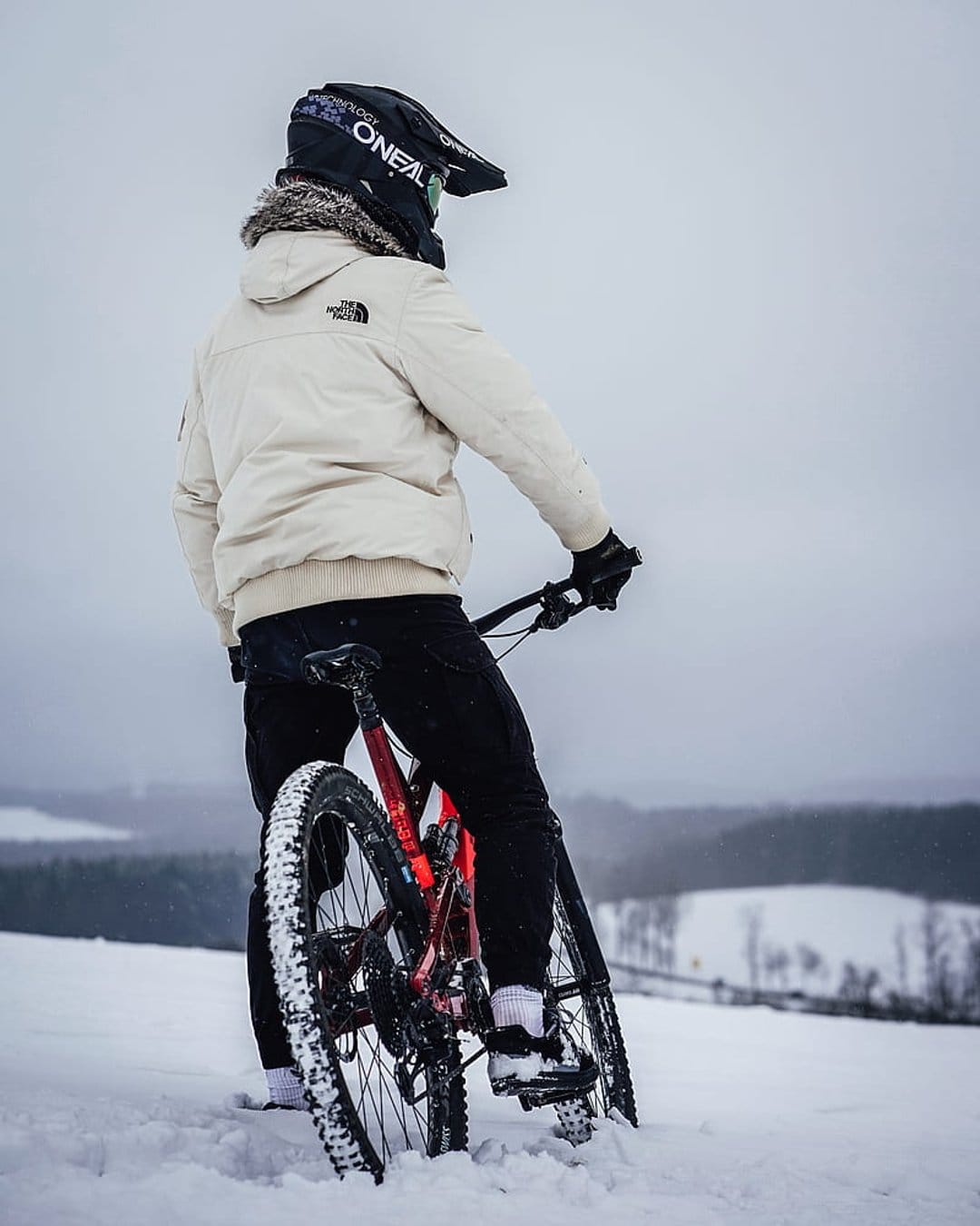Cyclist in a black helmet and cream jacket stands with a red mountain bike in deep snow amidst misty hills.